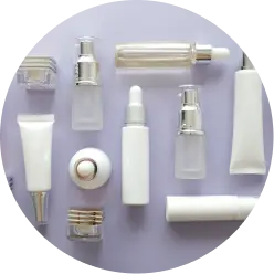 A flat lay image displays various clear and white cosmetic containers and bottles, including pump bottles, tubes, jars, and a dropper bottle, arranged neatly on a light purple surface. The containers are empty and ready for use.