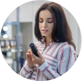 A woman with long dark hair, wearing a white and red striped shirt, is looking at a black bottle in her hand. She is standing in a store with shelves of products in the background.