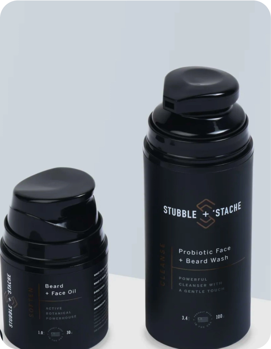 Two black cylindrical bottles with dispensers. The smaller bottle is labeled Beard + Face Oil, and the taller one is Probiotic Face + Beard Wash. Both have sleek, modern designs and the brand name Stubble + Stache.
